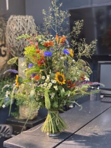 Wilder, natürlich gebundener Blumenstrauß in leuchtenden , Blau, gelb und orangeTönen mit vielfältigen Wiesenblumen, ergänzt durch filigrane Gräser und frisches Grün.