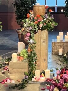 Urnenkranz aus pastell Blumen steht auf einer Säule und eine Urne steht im Kranz. Eine Ranke läuft vom Kranz an der Säule runter.