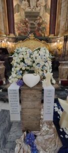 Engelsflügel aus weißen und hellblauen Blumen, davor liegt eine Urne in Herzform